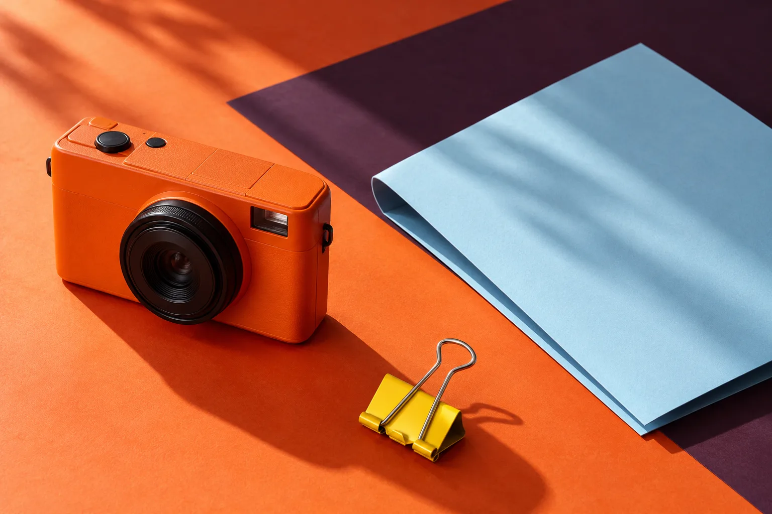 An unbranded orange camera, a folded blue brief, and a yellow clip on a studio tabletop